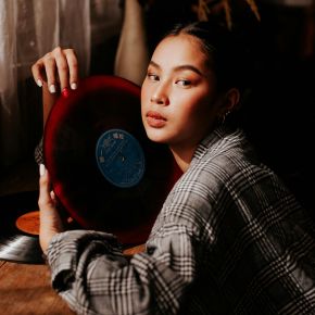Woman in plaid shirt holding a red vinyl record close to her face, sitting at a wooden table.