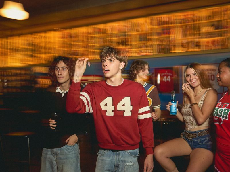 A group of young adults play darts and socialize in a lively bar or game room.