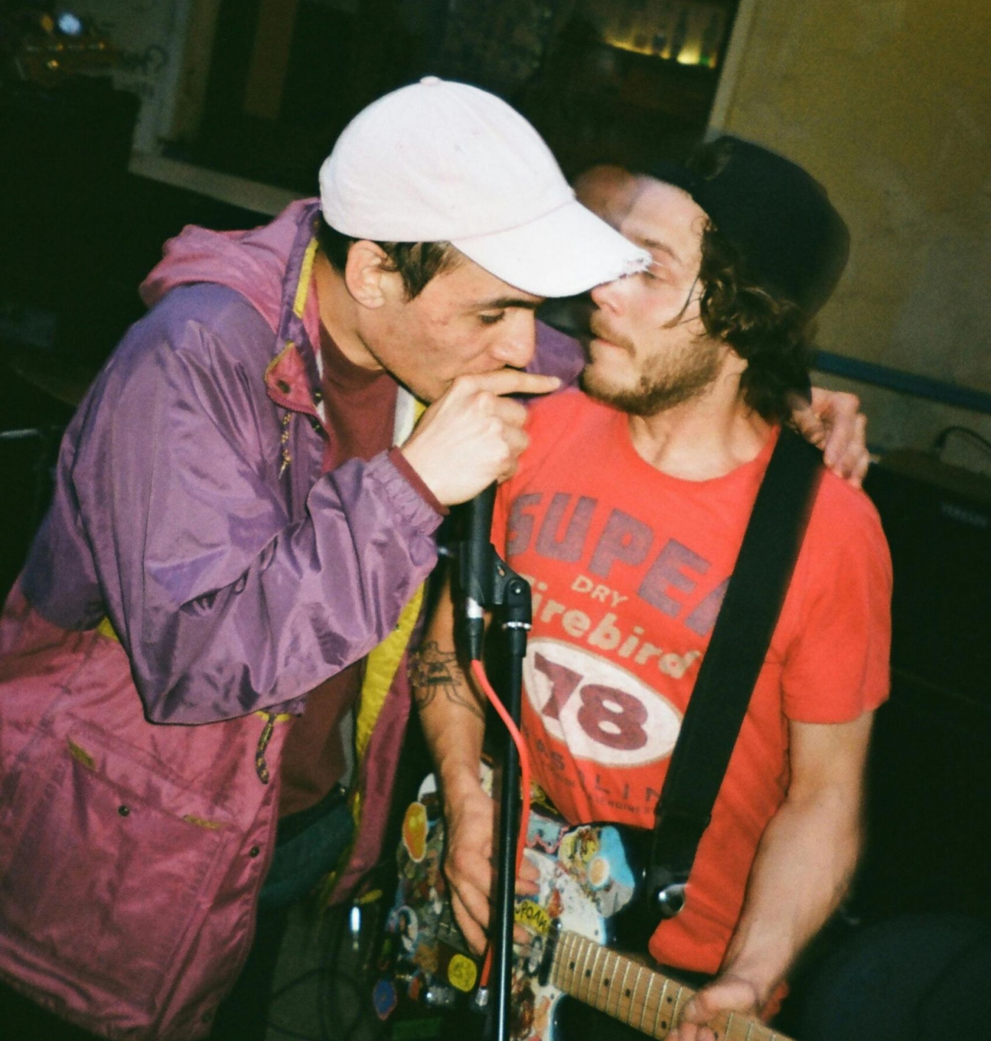 Two musicians perform closely together; one sings into a mic, the other plays a guitar covered in stickers.