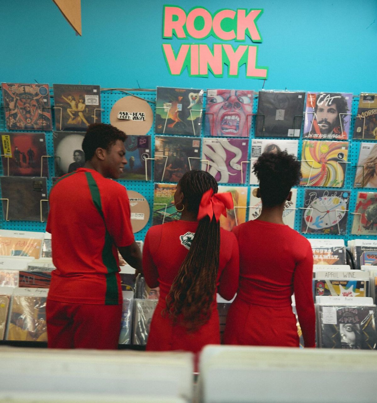Three people in red outfits browse rock vinyl records in a colorful record store with albums on the wall.