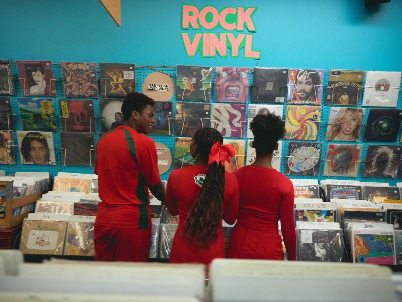 Three people in red outfits browse rock vinyl records in a colorful record store with albums on the wall.