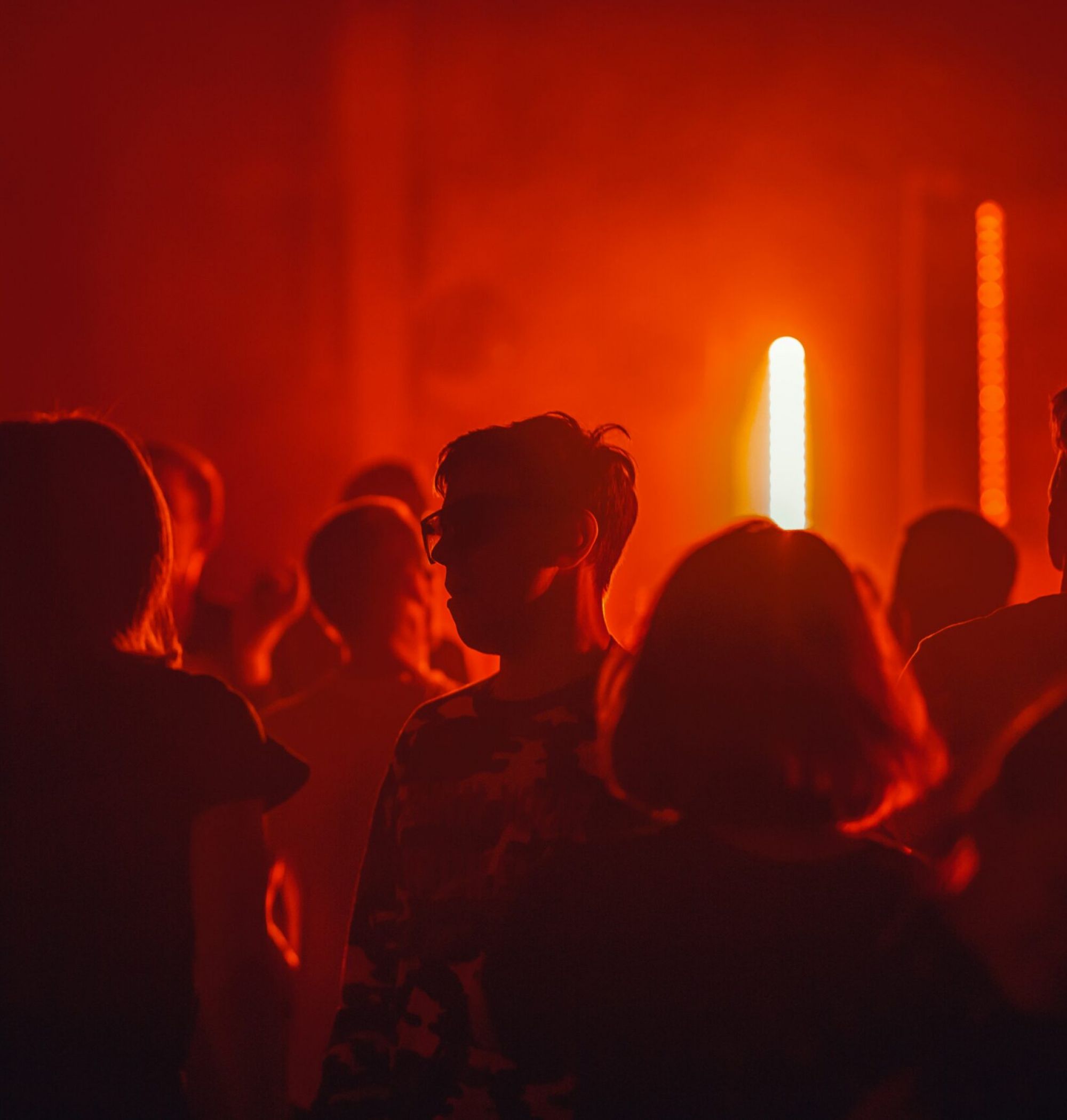 People dancing in a dimly lit room with red lighting and a bright light source in the background.