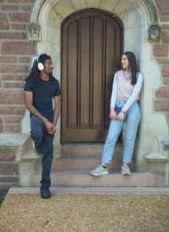 Two people smiling and talking while standing on steps in front of an arched wooden door.