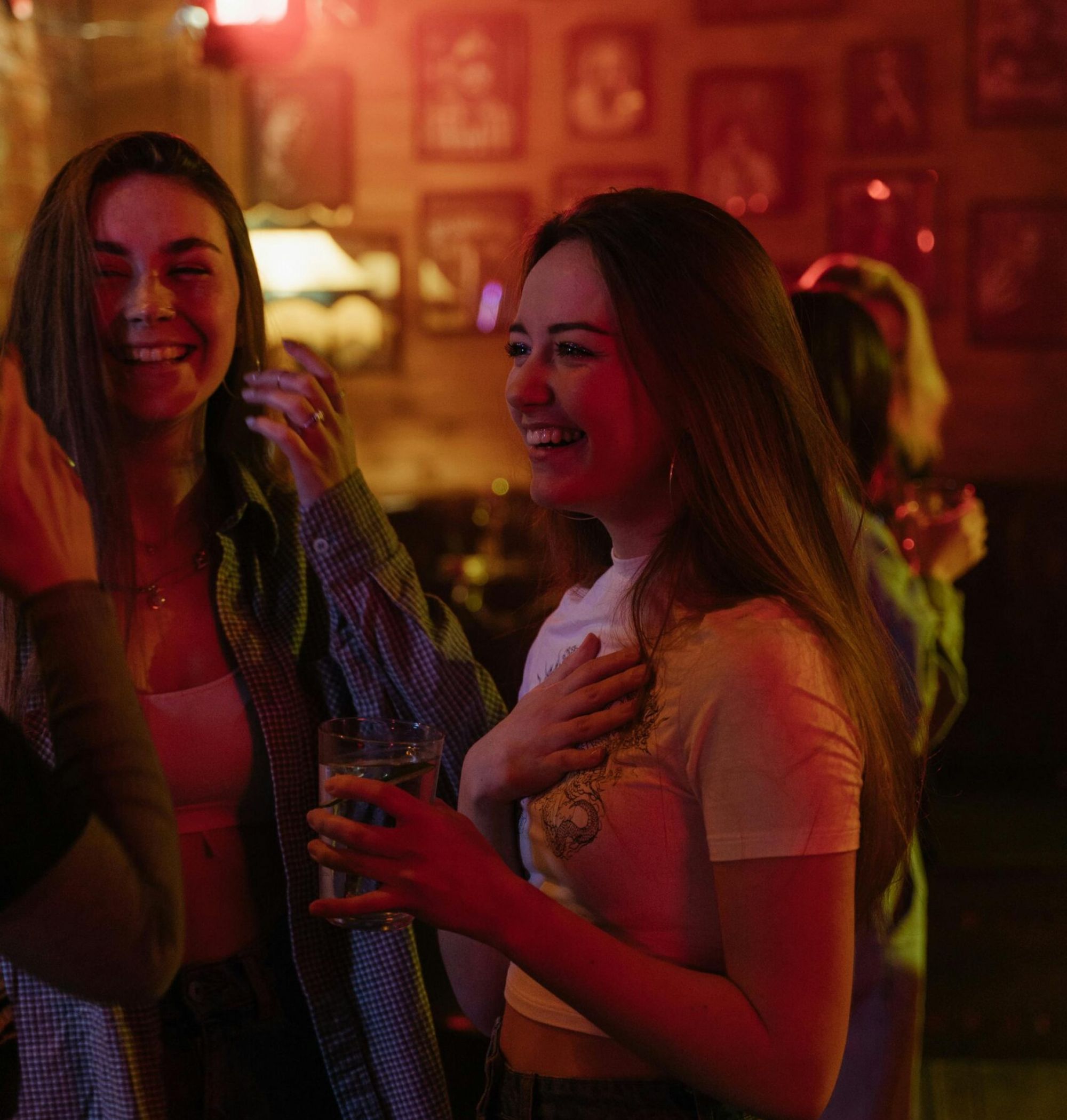 Two young women smiling and talking at a dimly lit bar with red lighting, holding drinks.