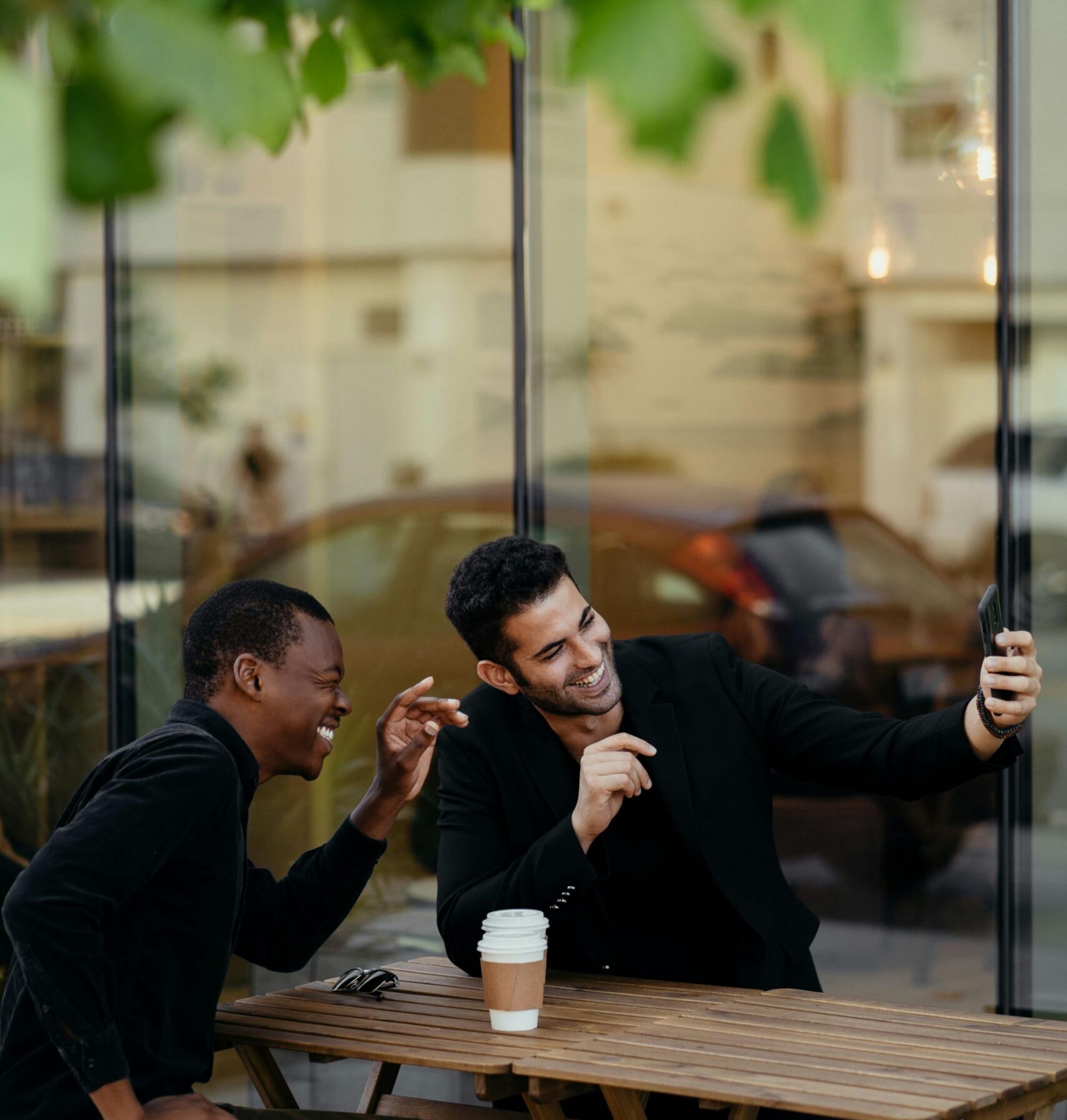 Two men sitting at an outdoor café table, laughing and taking a selfie together with a smartphone.