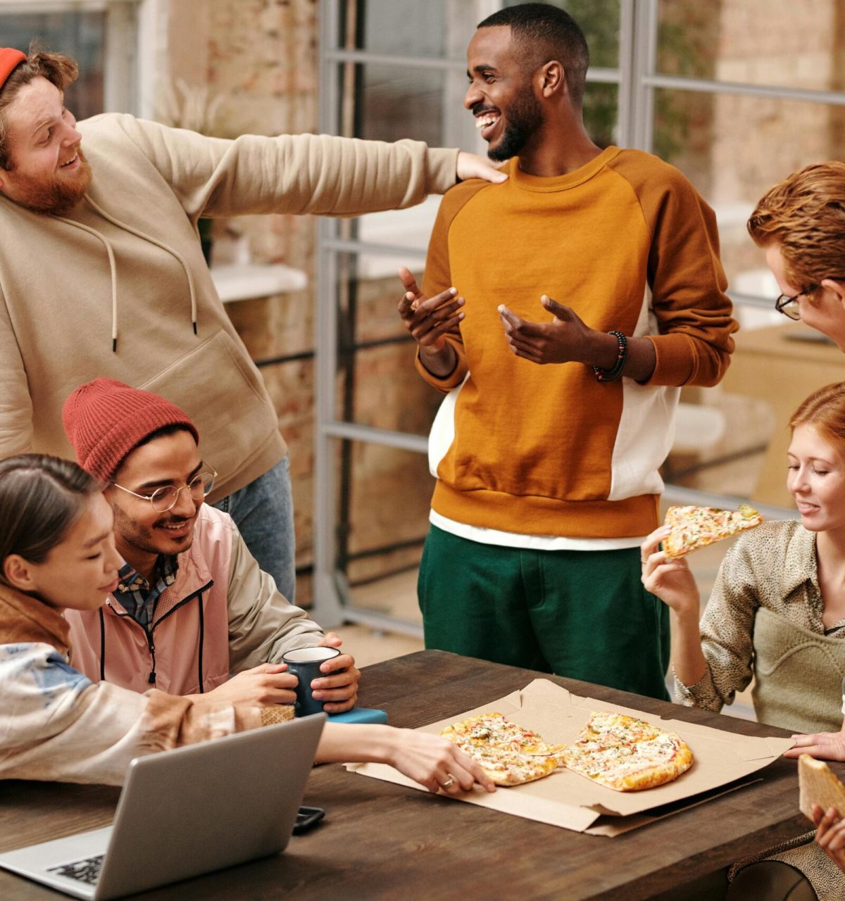 A group of friends laugh and eat pizza together around a table in a cozy indoor setting.