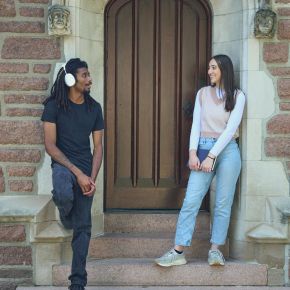 Two people smiling and talking while standing on steps in front of an arched wooden door.