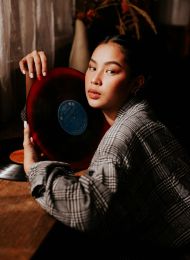 Woman in plaid shirt holding a red vinyl record close to her face, sitting at a wooden table.