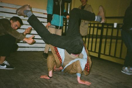 Person breakdancing indoors on wooden floor while others watch and gesture with thumbs up.