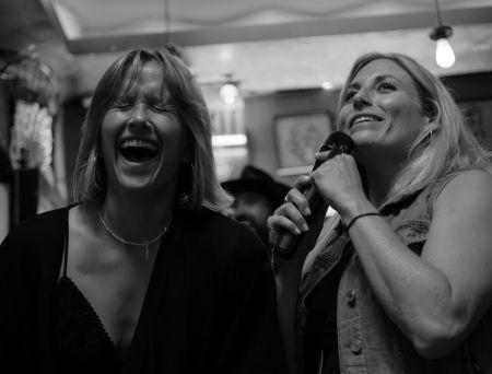Two women laughing and singing karaoke, one holding a microphone, in a lively indoor setting.