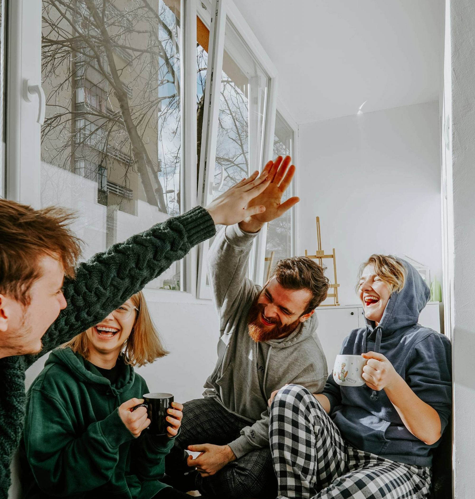 Four friends sit by a sunny window, laughing and giving high fives while drinking tea or coffee.