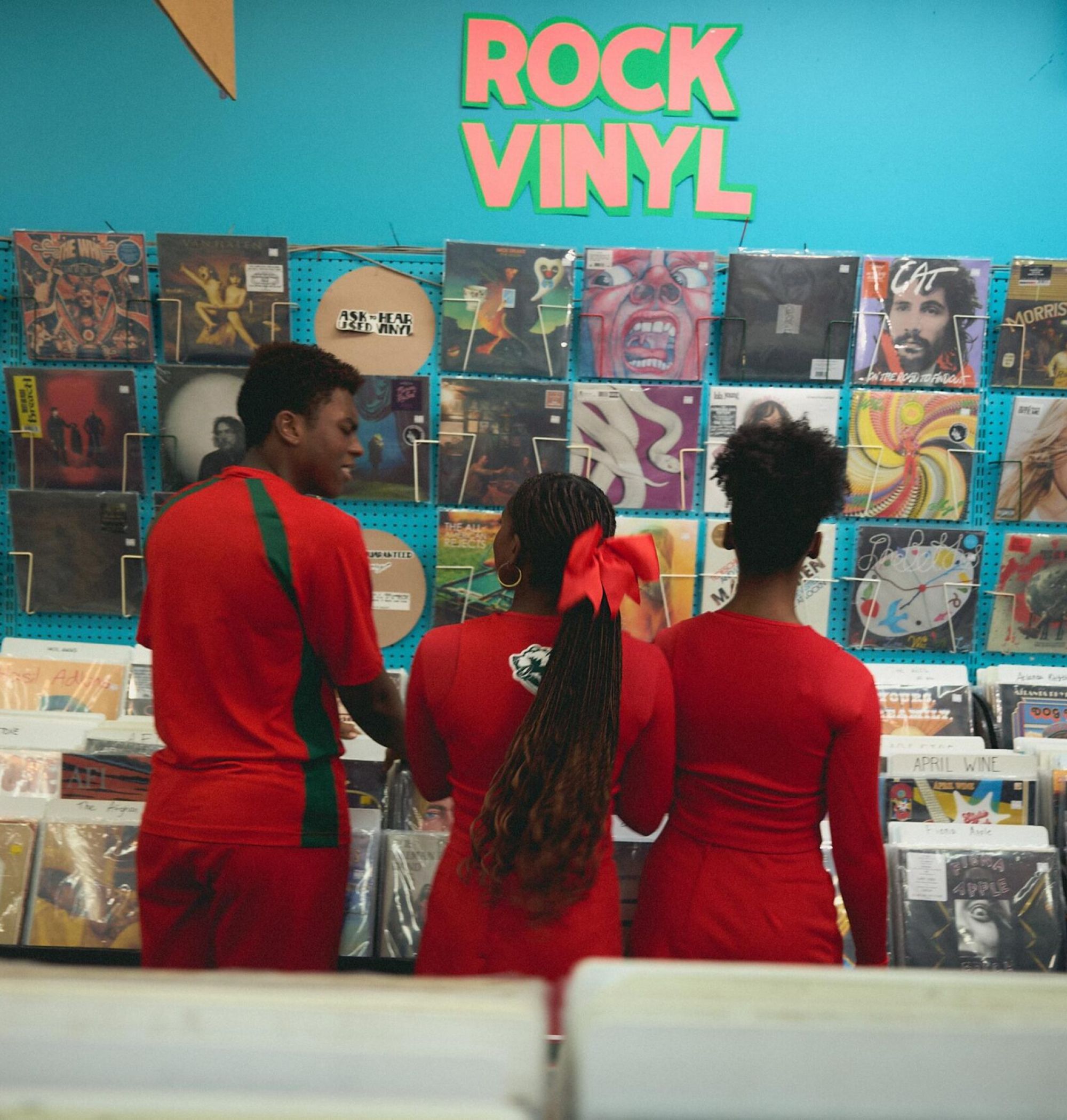 Three people in red outfits browse rock vinyl records in a colorful record store with albums on the wall.
