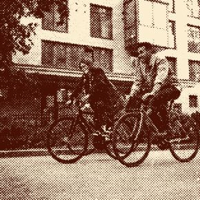 Two people ride bicycles side by side on a street in front of a modern apartment building.