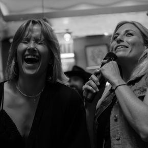 Two women laughing and singing karaoke, one holding a microphone, in a lively indoor setting.