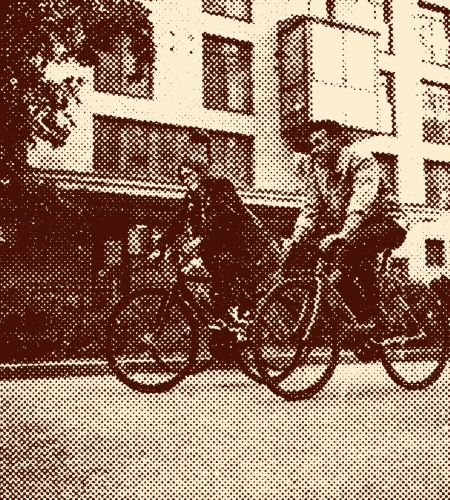 Two people ride bicycles side by side on a street in front of a modern apartment building.