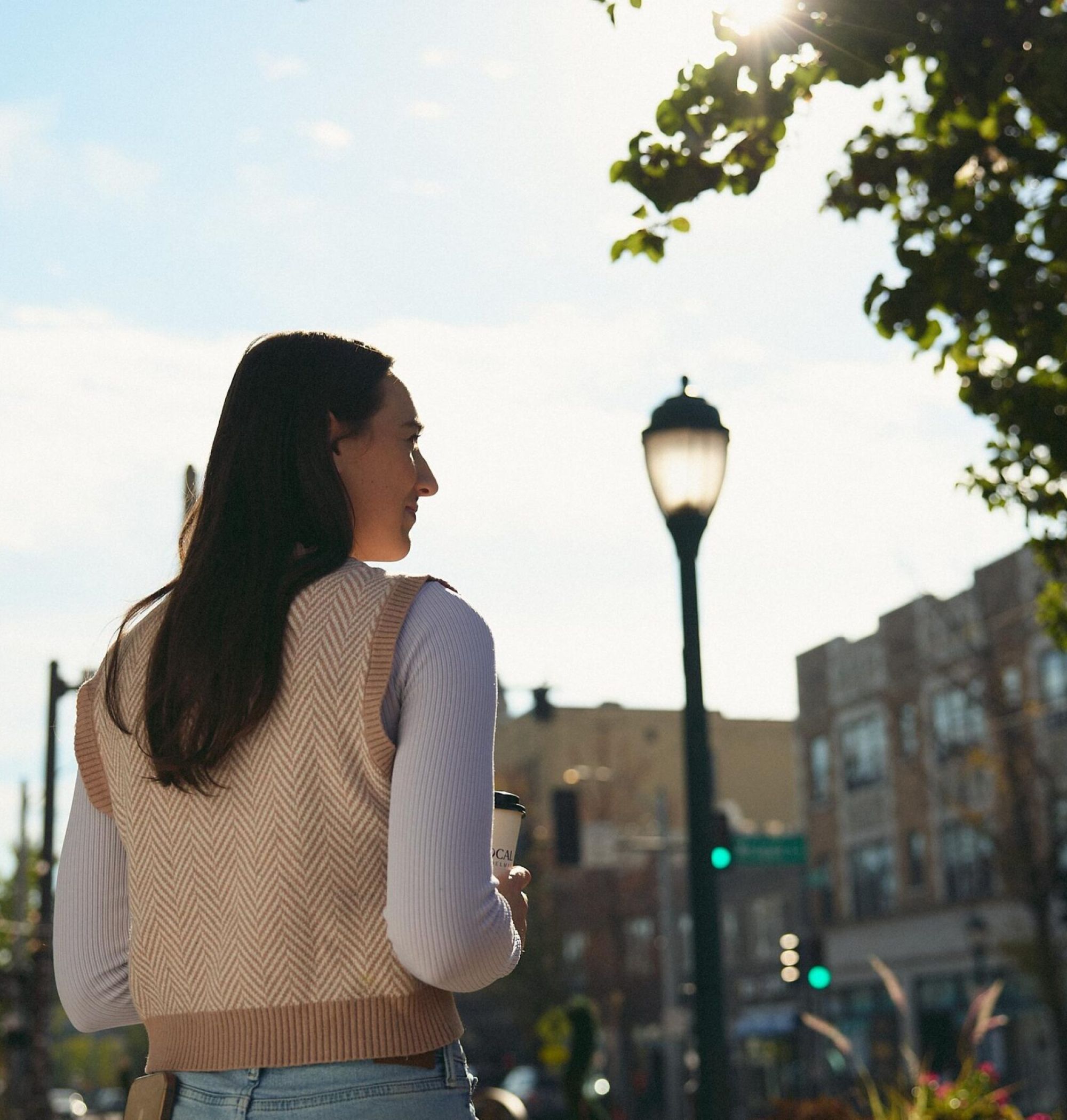 LOCAL On Delmar woman holding a cup walks outdoors on a sunny day with buildings and a streetlamp in the background.
