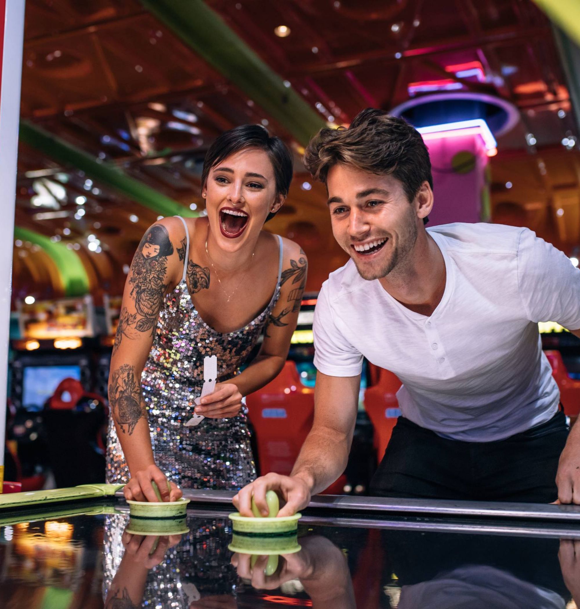 A smiling man and woman play air hockey together in a brightly lit arcade.
