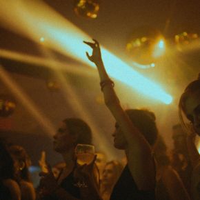 People dancing and raising drinks under golden lights at a lively, crowded nightclub or party.