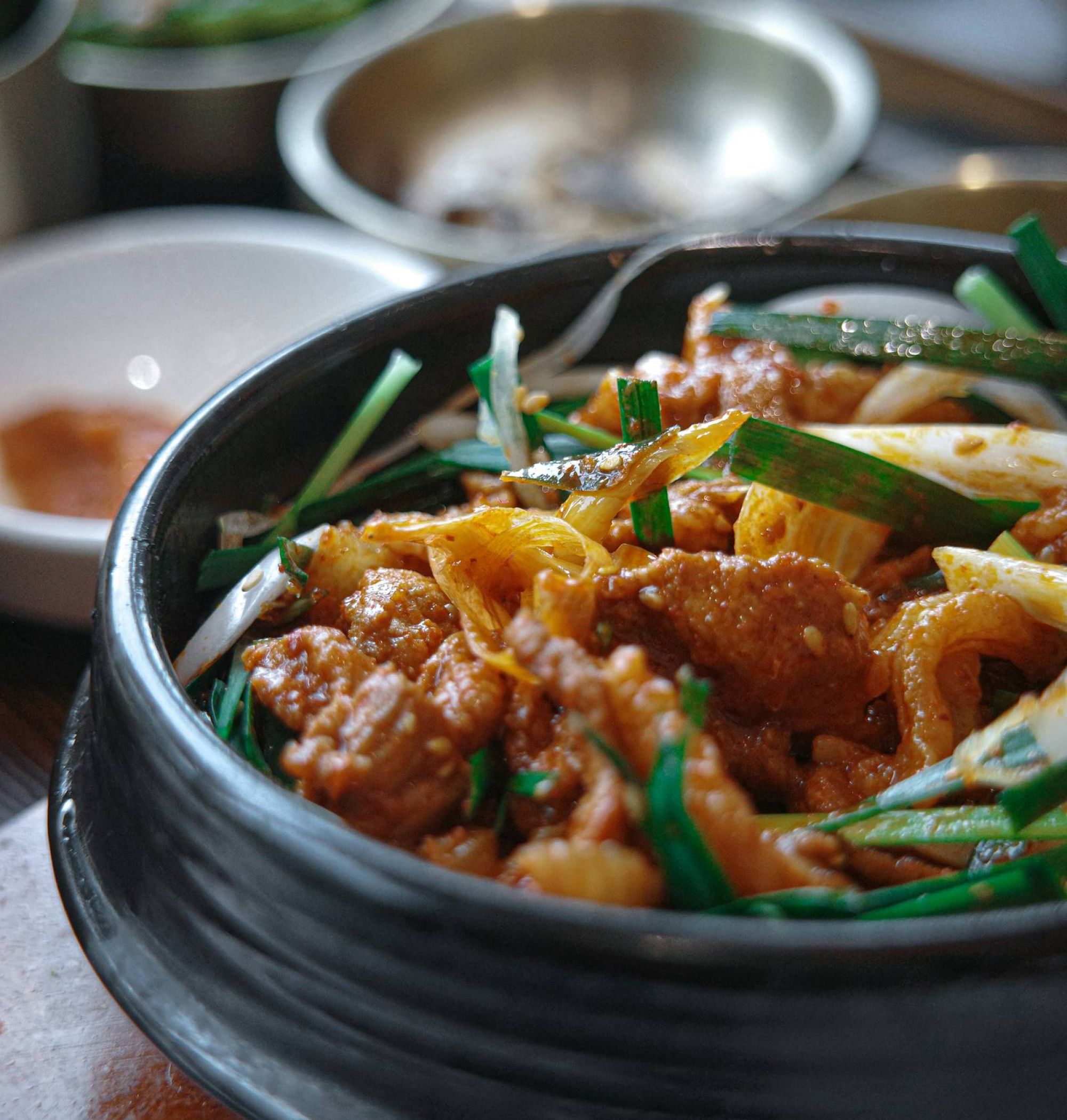 A bowl of stir-fried meat with vegetables, garnished with green onions, on a table with side dishes.
