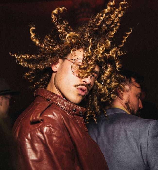 Person in a brown leather jacket with curly hair flying, dancing at a lively party.