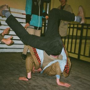 Person breakdancing indoors on wooden floor while others watch and gesture with thumbs up.