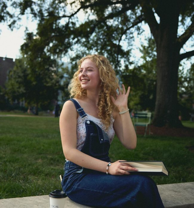 LOCAL On Delmar smiling woman in overalls sits on a bench outdoors, holding a book and waving, with a coffee cup nearby.