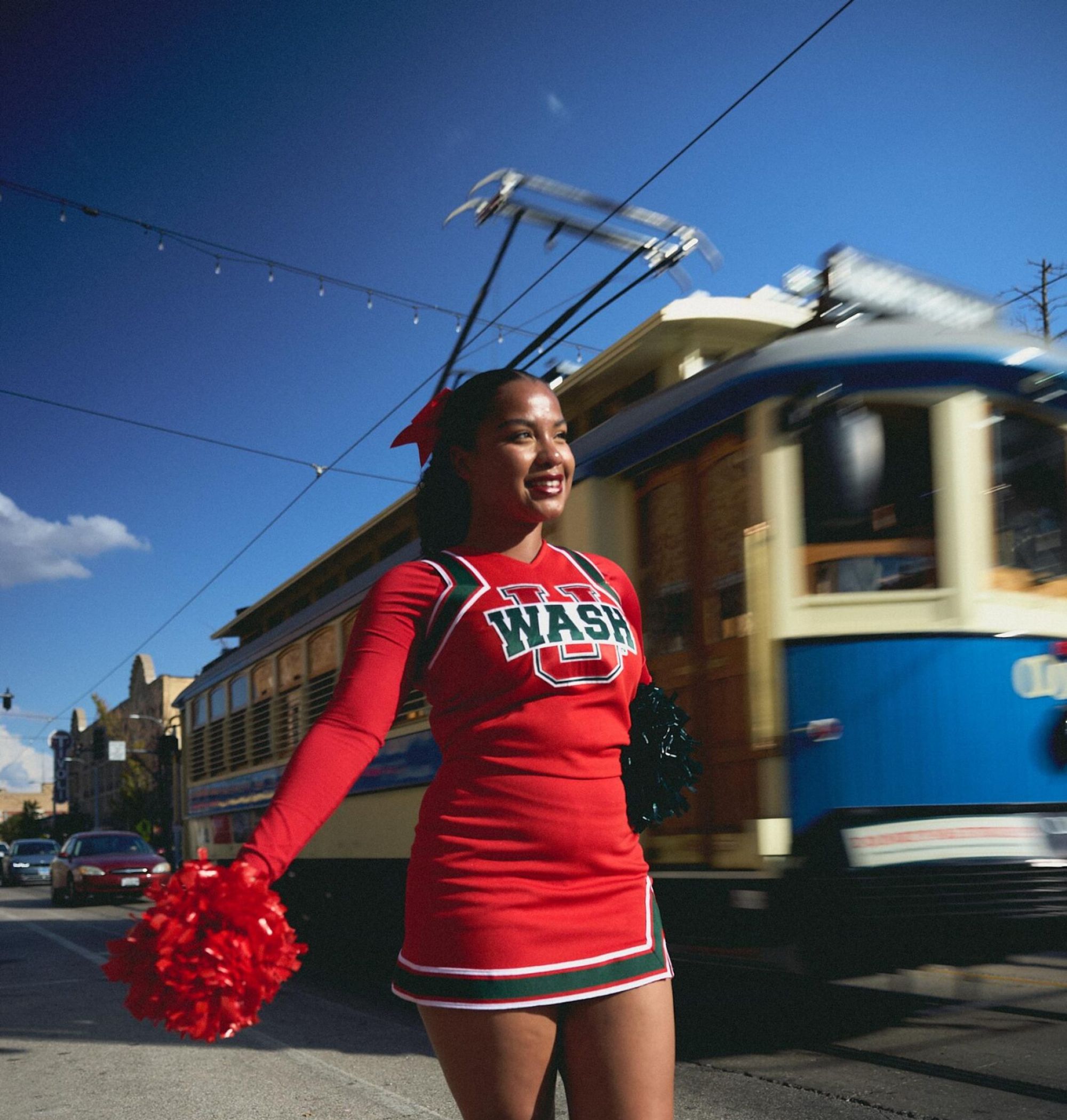A cheerleader in a red uniform poses on a street with a blue trolley passing by in the background.